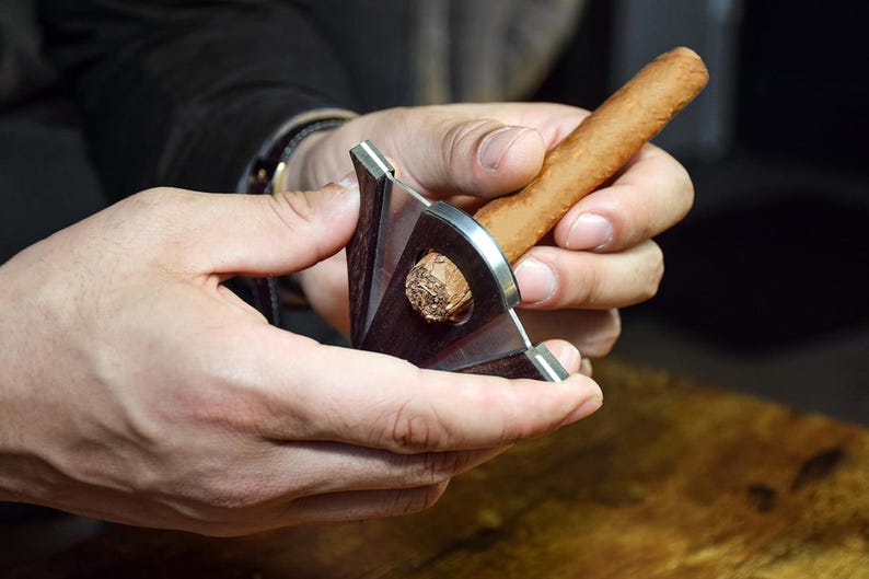 Puede incluir: Un cortapuros en V con cuerpo de madera marr&oacute;n oscuro y mecanismo de cuchilla de metal plateado. La herramienta est&aacute; colocada alrededor de un cigarro marr&oacute;n claro, mostrando la muesca en forma de V cortada en el extremo de la hoja de tabaco.