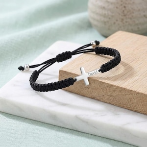 May include: A black cord bracelet featuring a silver-toned cross charm as the central element. The cross is engraved with the name David. The bracelet has an adjustable sliding knot closure with two small silver-toned beads at the ends of the cords.