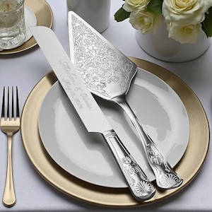 May include: Silver cake knife and server set with ornate floral detailing. The knife is engraved with the names "Michael & Olivia" and the date "May 15, 2026". The set is displayed on a white plate with a gold charger.
