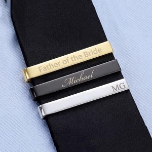 May include: Three rectangular tie clips in gold, black, and silver tones, each featuring custom engraving. The gold clip reads Father of the Bride, the black clip features the name Michael in script, and the silver clip displays the initials MG.