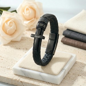 May include: Black leather bracelet with a woven design and a black cross charm. The cross has the name "Daniel" engraved on it. The bracelet is displayed on a white marble stand.