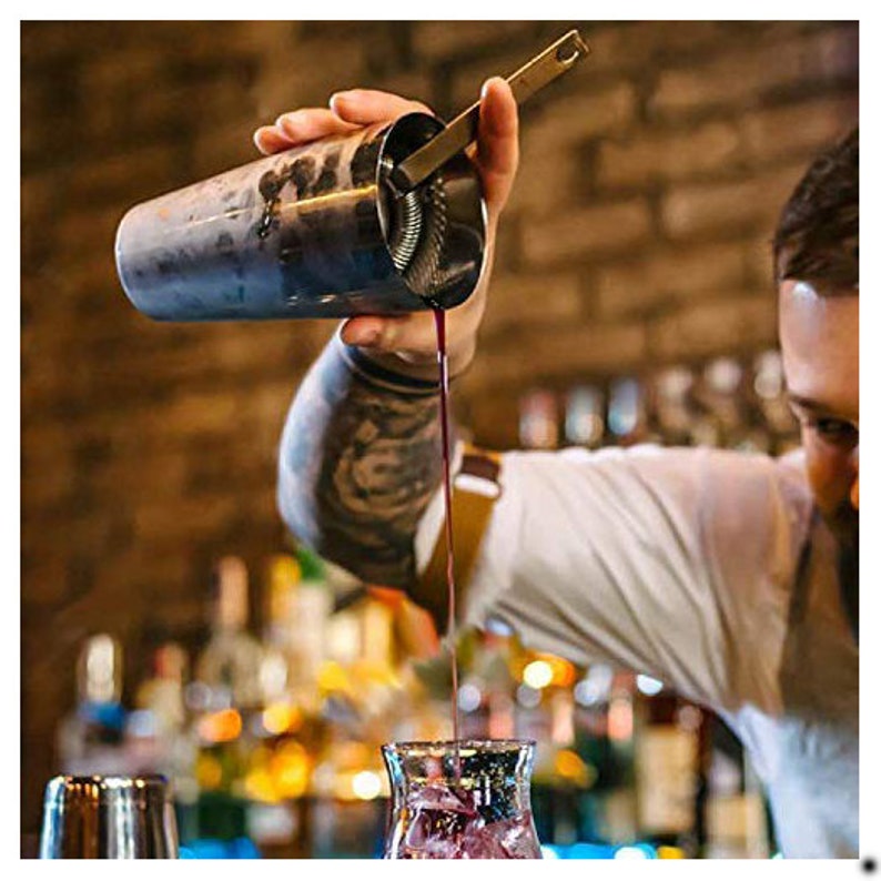 Op de afbeelding: Een barman giet een donkerrode vloeistof uit een cocktail shaker van roestvrij staal in een helder glas. De shaker heeft een ingebouwde zeef en een lange handgreep.