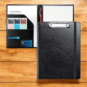 May include: Black leather clipboard folio with a pen holder, a card slot, and a lined notepad. The clipboard has a metal clip and a black elastic band. The folio is personalised with the name "Parker".