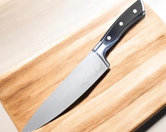 Puede incluir: Un cuchillo de chef plateado con mango negro y la inscripción "Chef Michael" en la hoja, descansando sobre una tabla de cortar de madera.