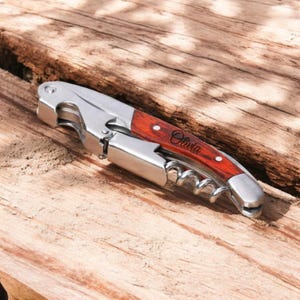 May include: A wooden and silver wine opener with a corkscrew and a bottle opener. The handle is engraved with the name "Olivia".