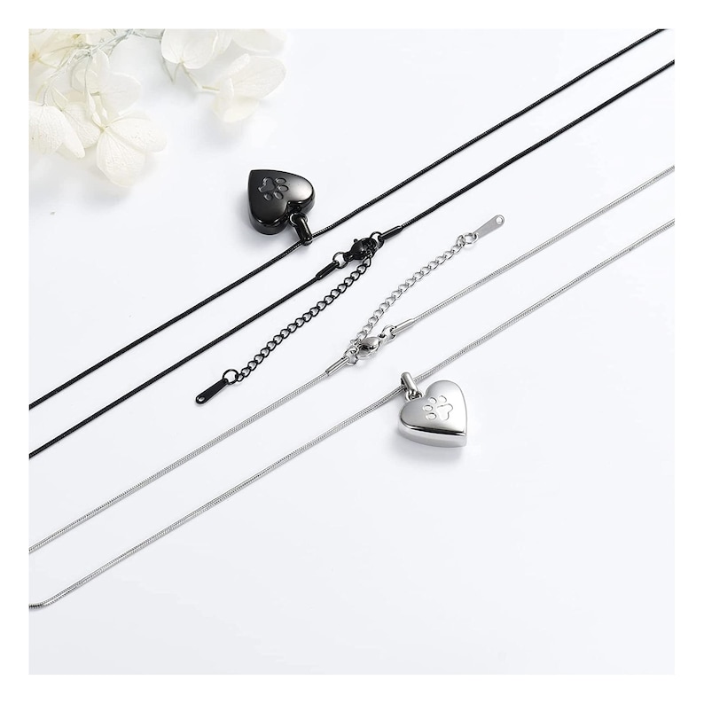 May include: Two silver heart-shaped cremation necklaces with a paw print design. One necklace has a black chain and the other has a silver chain.