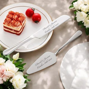 以下が含まれることがあります： 「David & Jessica October 20th 2022」と刻まれた銀製のケーキナイフとサーバーのセット。セットは、赤いベルベットケーキのスライスと2つの赤いトマトが載った白いプレートの上に置かれています。