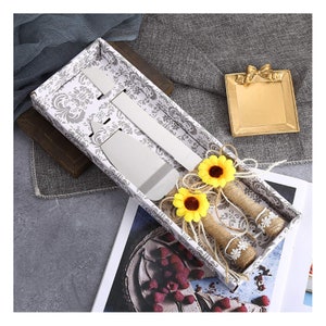 May include: A white box with a gray floral pattern contains two silver cake knives with sunflower accents. The knives are wrapped in twine and tied with a small sunflower.