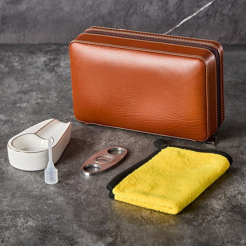 Op de afbeelding: Een bruine rechthoekige etui met ritssluiting, een witte keramische sigarenasbak met een gouden rand, een zilverkleurige metalen sigarenknipper, een kleine doorzichtige plastic druppelaar en een rechthoekige gele microvezeldoek.