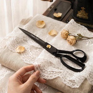 May include: Black metal scissors with a gold-toned pivot screw and the name Christina Smith engraved on the blade. The scissors feature two oval finger loops and are positioned on a piece of white lace fabric with dried rose flower heads.