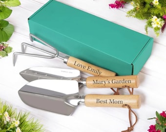 May include: A set of three gardening tools with wooden handles and personalized engravings. The tools include a trowel, a hand rake, and a small shovel. The tools are packaged in a teal box.