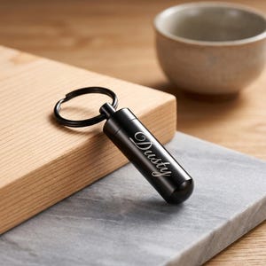 May include: A cylindrical black metal keychain urn featuring a screw-top lid and a circular split ring. The side of the black metal cylinder is engraved with the name Dusty in a white cursive script font, providing a clear contrast.