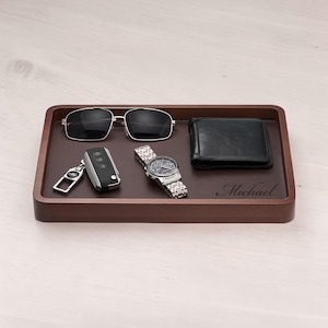 May include: A dark brown wooden tray with a leather-like interior holds sunglasses, a car key fob, a wristwatch, and a black leather wallet. The name "Michael" is engraved on the tray. The tray is rectangular.