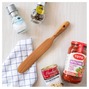 May include: A wooden spatula with the text "The Smiths" engraved on the handle. The spatula is on a white and blue checkered cloth. There are also various spices, sauces, and canned goods in the image.