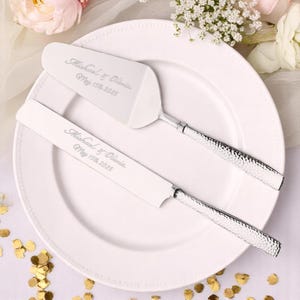 May include: A silver cake server and knife set with textured handles, resting on a white plate. The set is engraved with the names "Michael & Olivia" and the date "May 15th 2025". The background includes flowers and gold confetti.