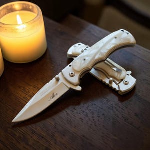 May include: A white folding knife with a pearlized handle and a silver blade. The blade is engraved with the name "Alex". The knife is lying on a dark wooden surface.