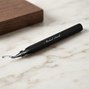 May include: A matte black craft knife with a hexagonal handle featuring the engraved name Michael Smith in a white script font. The tool has a silver-colored curved metal blade attached to the end of the sleek, elongated handle.
