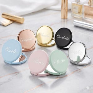 May include: A collection of personalized compact mirrors in various colors, including blue, pink, green, gold, rose gold, and black. Each mirror is round and features a name engraved on the front. The mirrors are open, revealing a reflective surface.
