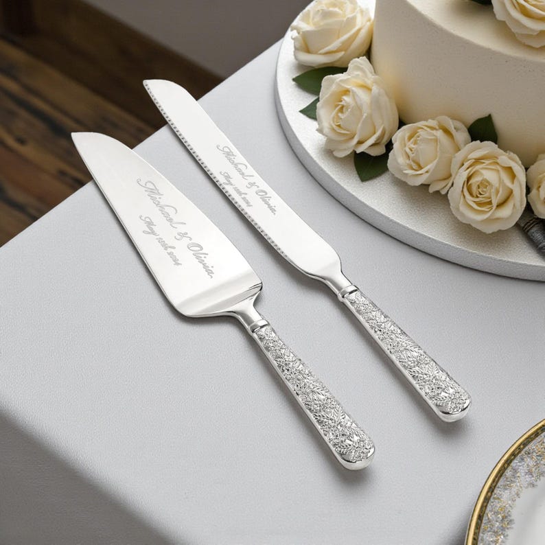 Puede incluir: Juego de cuchillo y servidor de pastel plateado con nombres y fecha grabados. Los mangos presentan un dise&ntilde;o floral detallado. El juego se exhibe cerca de un pastel blanco decorado con rosas blancas, ideal para una boda.