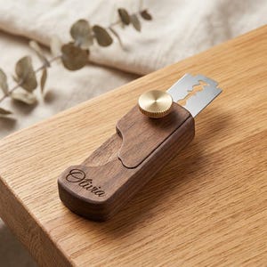 May include: A wooden bread lame with a dark brown finish, featuring the name Olivia engraved in script on the handle. The tool includes a silver-coloured metal razor blade secured by a circular, textured brass-coloured knob on the top surface.