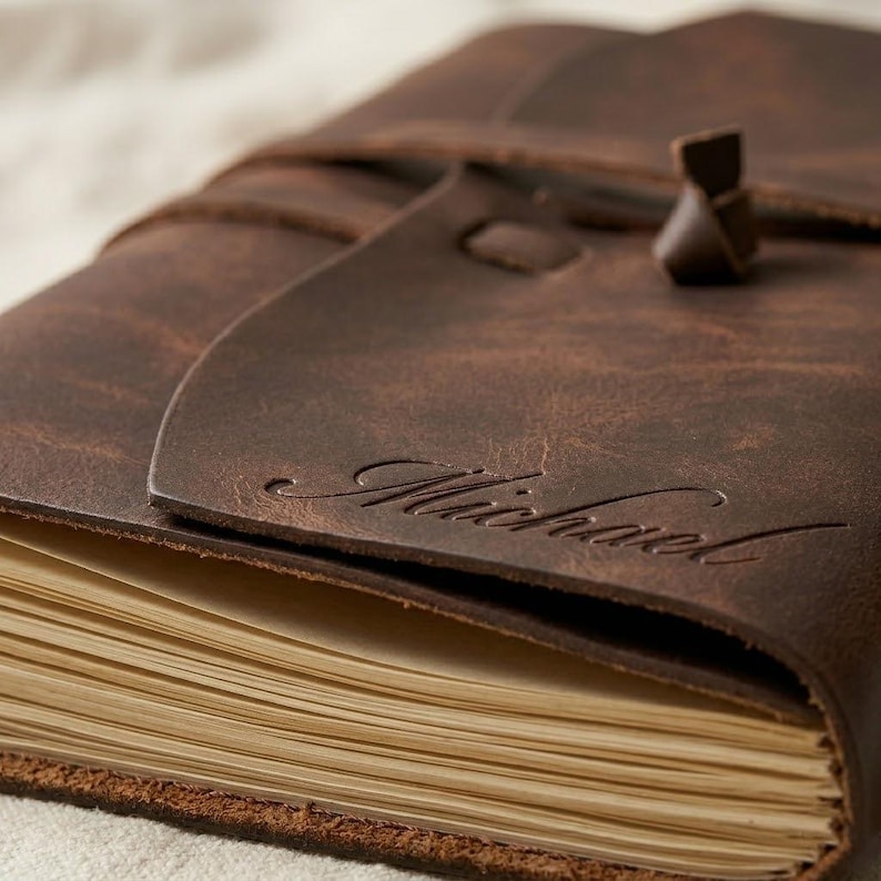 May include: A dark brown leather journal with a wrap-around strap closure. The front flap features the name Michael embossed in a cursive script. The journal contains numerous cream-colored pages visible along the side edge of the binding.