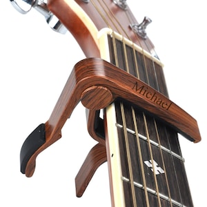 Op de afbeelding: Een bruine houten gitaarkapodaster met een zwarte rubberen grip en de naam "Michael" gegraveerd aan de zijkant. De kapodaster is bevestigd aan de hals van een akoestische gitaar.