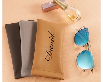Peut inclure: Trois étuis à lunettes de différentes couleurs, un marron, un gris et un beige. L'étui beige porte le nom "David" écrit en cursive noire. Une paire de lunettes de soleil bleues miroir et un flacon de parfum rose et or sont également présents sur l'image.