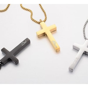 Könnte beinhalten: Drei silberne Halsketten mit verschiedenen Gravuren. Das Kreuz links ist schwarz und mit dem Wort "Dad" graviert. Das Kreuz in der Mitte ist golden und mit dem Wort "Alice" graviert. Das Kreuz rechts ist silber und mit römischen Ziffern graviert.