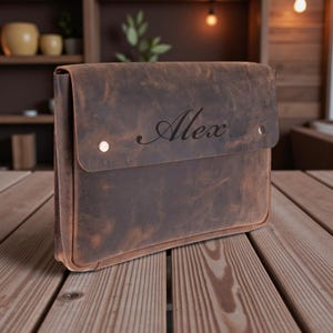 May include: A brown leather laptop case with a flap closure and two silver rivets. The name "Alex" is elegantly scripted on the front. The case is made of distressed leather, giving it a rustic appearance.