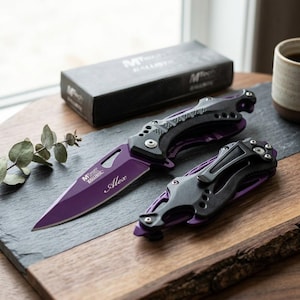 May include: A folding pocket knife with a purple blade featuring the name Alex and the MTech Ballistic logo, paired with a black handle. A second, closed folding knife with a black handle and purple accents lies beside it, along with a black box.