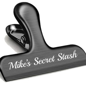 Puede incluir: Un clip de metal negro con acabado cepillado y el texto "Mike's Secret Stash" grabado en la parte delantera.