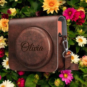 May include: A brown leather camera case with a strap and the name "Olivia" engraved on the front.