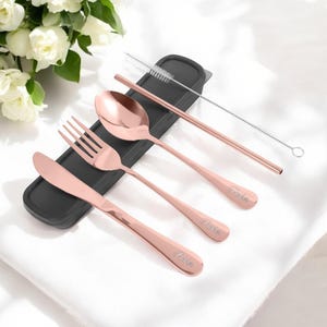 May include: A set of rose gold cutlery with a straw and cleaning brush. The cutlery includes a knife, fork, spoon, and straw. The set is packaged in a black case with the word "Paris" engraved on each piece.