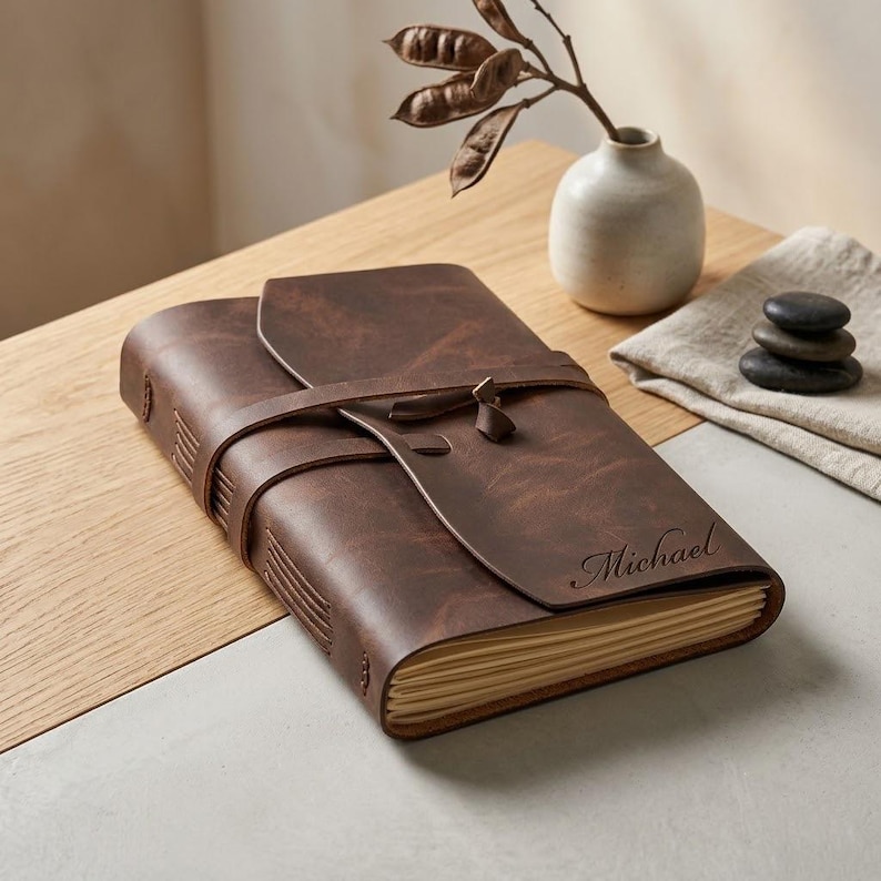 May include: A dark brown leather journal with a wrap-around strap closure and visible stitching along the spine. The front cover features the name Michael engraved in a cursive script. The journal contains cream-colored pages with deckled edges.