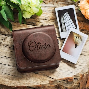 May include: A brown leather camera case with a circular cutout on the front. The case has the name "Olivia" engraved on the front.
