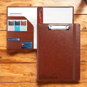 May include: A brown leather clipboard folio with a pen, notepad, and card slots. The folio has a silver clip and an elastic band closure. The name "Parker" is engraved on the front. The folio is open, revealing a white notepad and a pen.