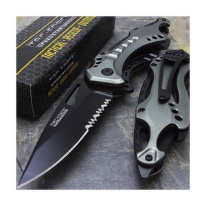 May include: A black and gray tactical folding knife with a serrated blade and a bottle opener. The knife has a black handle with a textured grip and a gray metal frame. The knife is open and the blade is extended. The knife is on a gray surface.