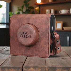 Op de afbeelding: Een bruine leren camerahoes met de naam "Alex" gegraveerd op de voorkant. De hoes heeft een getextureerde afwerking en een bijpassende bruine riem. De camerahoes staat op een houten oppervlak.