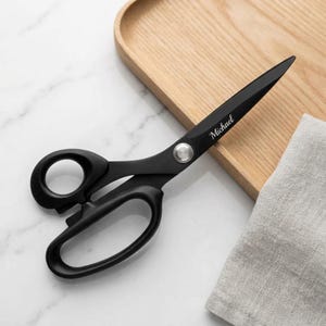 Puede incluir: Tijeras de color negro mate con un tornillo central plateado y el nombre Michael grabado en una fuente cursiva blanca en la hoja superior. Las tijeras cuentan con dos anillos para los dedos diferenciados y una punta afilada para cortar.