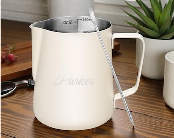 May include: Cream-colored milk frothing pitcher with a stainless steel interior and handle. The pitcher has a capacity of 12 oz. and features the name "Parker" in a script font. A stainless steel stirring spoon is inside the pitcher.
