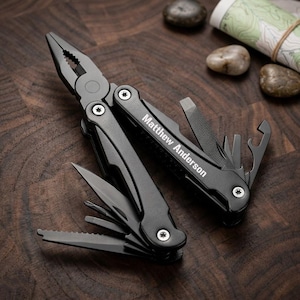 May include: A black multi-tool with various implements, including pliers, a knife, and a bottle opener. The tool is personalized with the name "Matthew Anderson". The tool is open, displaying its different functions. The background includes rocks and a rolled-up map.