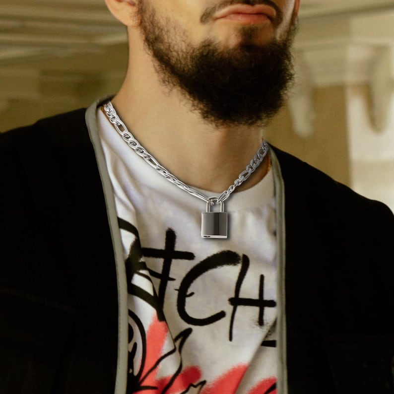 May include: A silver chain necklace with a silver padlock pendant. The necklace is worn by a person with a beard.