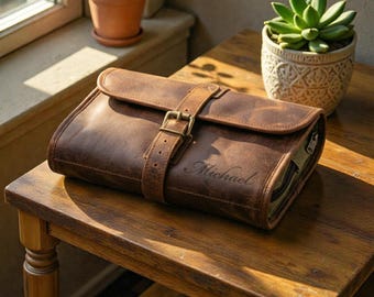 Peut inclure: Une trousse de toilette en cuir marron avec une fermeture à boucle et le nom "Michael" gravé sur le devant. Le sac est posé sur une table en bois. Le sac est conçu pour contenir des articles de toilette et autres effets personnels.