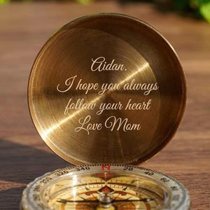 Puede incluir: Una brújula de latón con una esfera detallada y una tapa grabada con las palabras "Aidan. Espero que siempre sigas tu corazón Love Mom." La brújula está abierta, revelando la inscripción y el dial direccional.