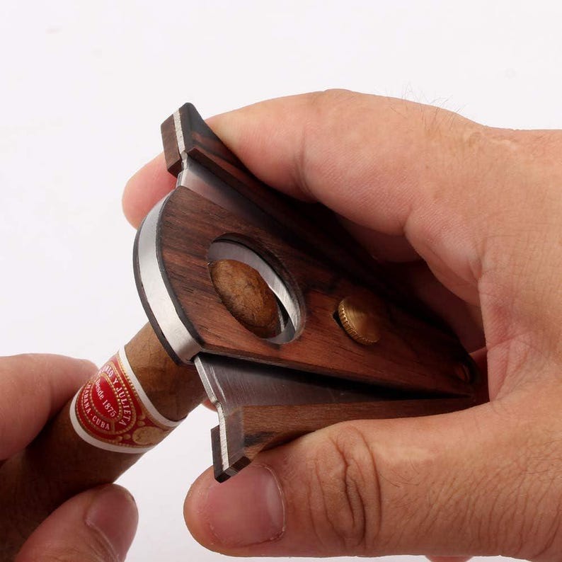 Puede incluir: Un cortapuros con acabado de veta de madera marr&oacute;n oscuro, una hoja de metal plateado y una abertura circular. El cortador tiene un peque&ntilde;o detalle de tornillo de color lat&oacute;n en el lateral y est&aacute; colocado alrededor de un cigarro marr&oacute;n con una anilla roja y blanca.