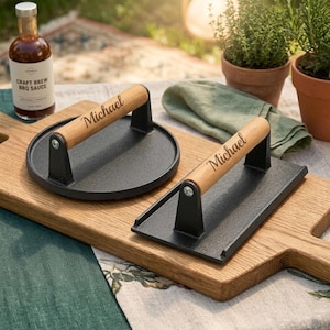 May include: Two cast iron grill presses with wooden handles, one round and one rectangular, each engraved with the name "Michael." A bottle of BBQ sauce and potted herbs are in the background. The presses sit on a wooden cutting board.