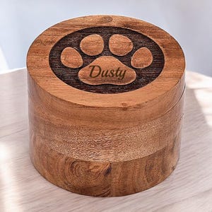 Op de afbeelding: Een houten doos met een poot afdruk ontwerp en de naam "Dusty" erop gegraveerd. De doos is bruin en heeft een gladde afwerking.