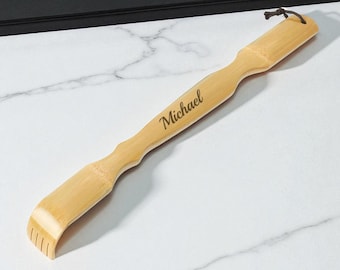 May include: A light brown bamboo back scratcher with the name "Michael" engraved on the handle. The scratcher has a curved head with vertical tines and a loop for hanging. The product is made of bamboo.