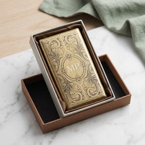 May include: A rectangular gold-toned metal case featuring an intricate engraved scrollwork pattern and the letters MP centered in an oval frame. The case is presented inside a brown box with a white inner tray and dark brown fabric lining.