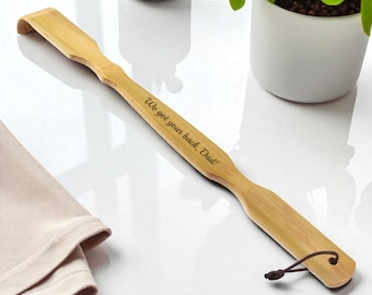 May include: A light brown bamboo back scratcher with the engraved text "We got your back, Dad!" A small brown cord is attached to the end. The back scratcher is on a white surface, with a small plant and a white container in the background.